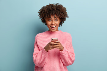 Photo of delighted beautiful female holds mobile phone, types text message, connected to wireless...