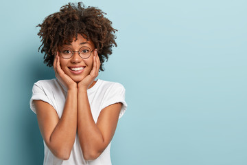 Isolated shot of good looking positive Afro female holds both hands on face, smiles happily,...