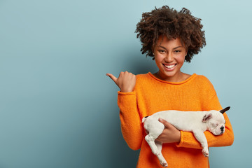 Horizontal shot of pleased Afro woman points away at free space, shows direction to pet store,...