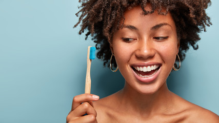 Headshot of curly delighted woman has dark skin, cleans teeth with toothbrush and paste, poses half...