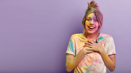 Impressed touched lady keeps both hands on chest, satisfied having Indian festival of colors, dirty with colored dye powder, stands over purple studio wall, free space on left for your promotion