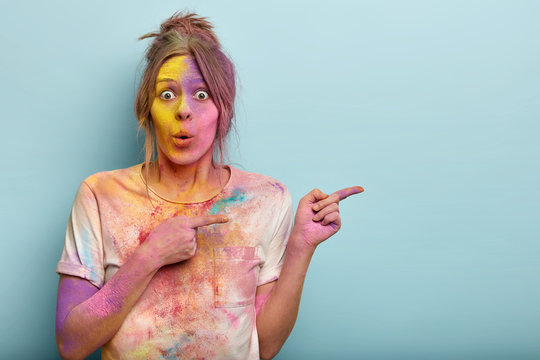 Studio Shot Of Surprised Emotional Female Model Has Bated Breath, Dirty With Colorful Powder, Has Multicolored Face, Demonstrates Something On Blank Space. Holi Festival Celebration Concept.