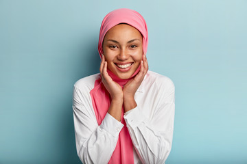 Headshot of pleasant looking Muslim woman touches cheeks with both hands, shows white teeth, wears...