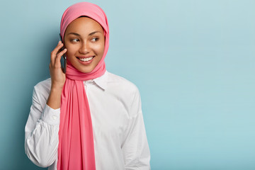 Isolated shot of pleasant looking female model speaks over modern mobile phone, talks with someone, smiles sincerely, focused away, stands against blue wall with empty space, wears Muslim clothes