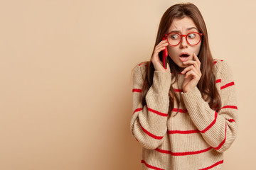 Puzzled beautiful female feels embarrassed, makes telephone conversation, uses application on cell...