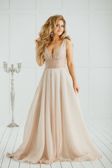 Fototapeta premium Beautiful blonde woman with greek hairstyle in beige powdery atlas wedding dress posing in studio room.