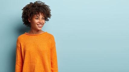 Image of dreamy woman with crisp hair looks pensively aside, has gentle smile on face, wears orange...