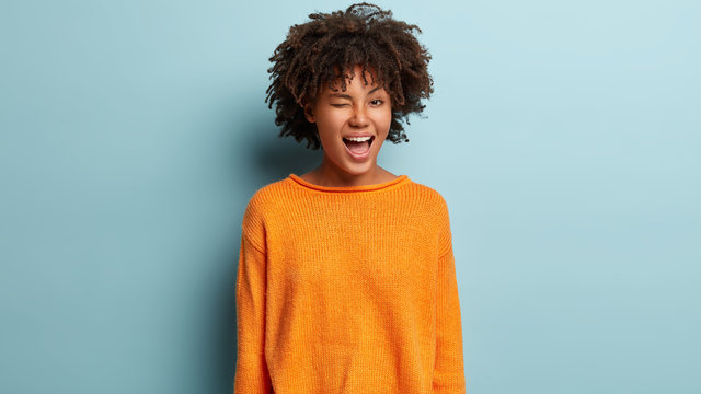 Portrait of playful funny young woman with glad expression, blinks eye, flirts with boyfriend, expresses good emotions, wears orange jumper, isolated over blue background. Hey, guy look at me.