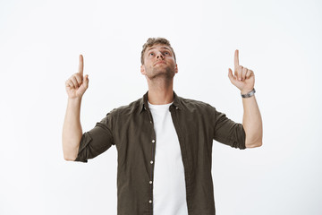 Curious and dreamy good-looking intrigued european blond guy with bristle in watch and casual outfit raising hand looking and pointing up using index fingers, focused on upwards copy space