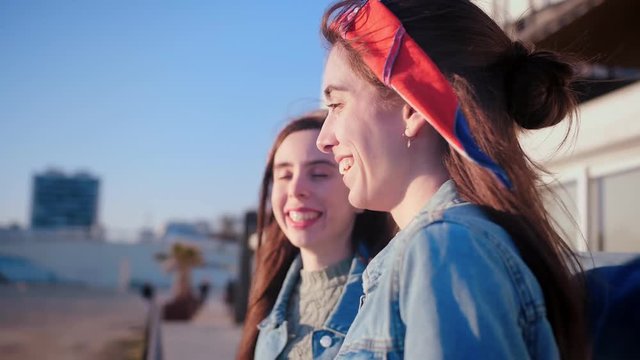 Portrait of young emotional Hispanic girls, share happy moments together, spend free leisure time with friend, gently hug and enjoy summer urban view, dressed in jeans jacket and colourful headband