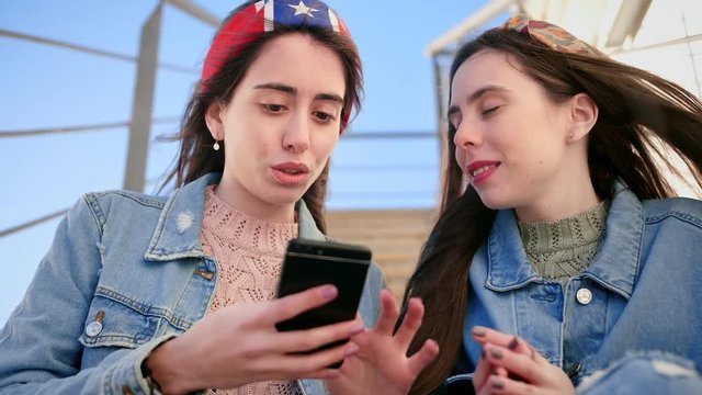 Two nice girl friends hanging out at stairs, using mobile phone sending text message Snapchat sharing digital content on social media enjoying free time, dressed jeans jacket and colourful headbands