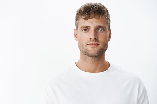 Waist-up shot of handsome calm blond blue-eyed guy with bristle in white t-shirt looking at camera with relaxed carefree facial expression posing over gray background, looking sincere and chill