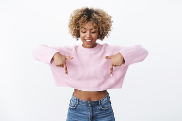 Curious and stylish fashionable african-american girl with blond curly haircut looking interested with broad smile as pointing down intrigued with cool amazing promotion over white background © Cookie Studio