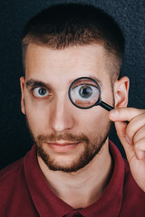 Fototapeta na wymiar a young man with a beard looks through a magnifying glass. Portrait of a guy with a big eye on a black background. investigation, survey