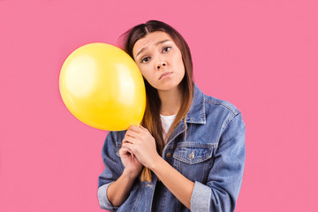 Sad girl holding a yellow balloon with a picture of a smiling face on an isolated pink background.