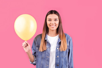 brunette girl in denim jacket holding a yellow balloon with a smiling face. Happy girl holding a balloon on a stand-pink background