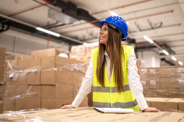 Hardworking professional female worker or manager with hardhat and reflective jacket leaned on cardboard boxes looking aside in large warehouse.