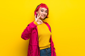 Young woman with pink hair over yellow wall showing ok sign with fingers