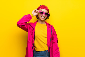 Young woman with pink hair over yellow wall with glasses and happy