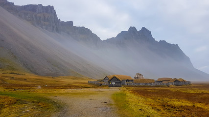 A small abandoned vikings village. Sod rooftops, turf rooftops. Village located at the bottom of a high mountain. Around the farm a wooden fence. Dry grass all around. Tall mountains being a shelter.