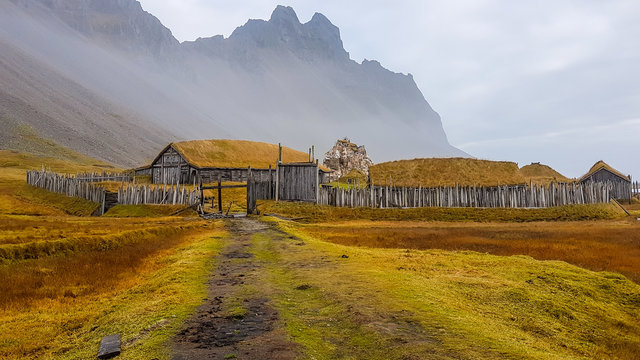 An abandoned vikings village. Sod rooftops, turf rooftops. Village located at the bottom of a high mountain. Around the farm a wooden fence. Dry grass all around. Traditional Scandinavian architecture