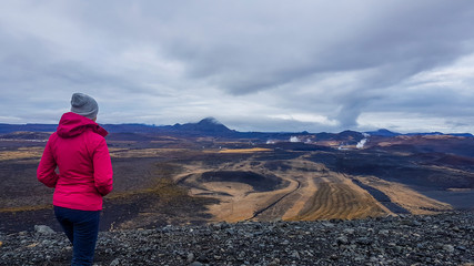 Fototapeta na wymiar A girl standing on a rocky surface of a volcano, overlooking a geothermal activity region. Girl wears pink jacket. Foam is coming out of the ground. Geothermal energy. Steam power.