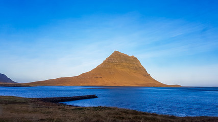 Fototapeta na wymiar A stunning view on famous Iceland's mountain, Kirkjufell. In front of the tall hat-like mountain spreads the shallow water of a fjord. Beautiful and clear day, popular destination for a holiday.