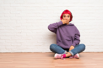 Young woman with pink hair sitting on the floor covering mouth with hands