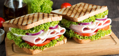Close-up photo of a club sandwich. Sandwich with meat, prosciutto, salami, salad, vegetables, lettuce, tomato, onion and mustard on a fresh sliced rye bread on wooden background. Olives background.