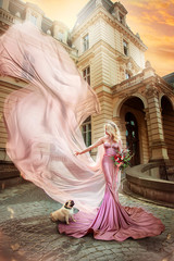 Fototapeta na wymiar Princess in pink dress with long veil on background of building at sunset