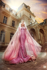 Princess blonde in pink dress with long shawl on background of building at sunset