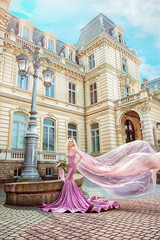 Princess blonde in pink dress on background of building at sunset. Wind raises shawl © popovatetiana