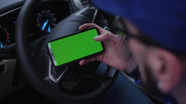 Young man sitting in driver's seat, holding smartphone in right hand, touching screen. Guy messaging with friend. App. Without face.