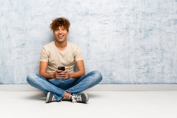 Young african american man sitting on the floor sending a message with the mobile