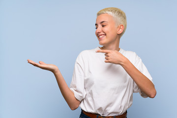 Teenager girl with white short hair over blue wall holding copyspace imaginary on the palm to...