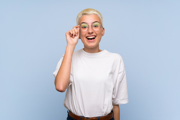 Teenager girl with white short hair over blue wall with glasses and surprised