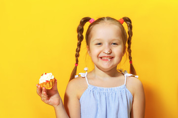 Closeup portrait of a cheerful little sweet tooth girl on a yellow background. The child smeared his nose with cream and holding a cake in his hands.