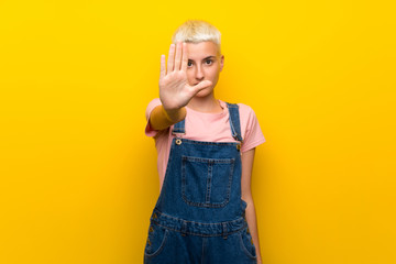 Teenager girl with overalls on yellow background making stop gesture