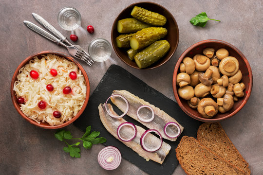 Traditional Russian snacks and vodka, sauerkraut with cranberries, herring, pickled cucumbers, pickled mushrooms and rye bread on a brown background. Top view