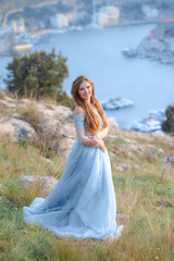 Fototapeta na wymiar A girl with long hair on the beach. She's wearing a beautiful blue dress . In the background view of the Bay, sea and city. Soft focus.