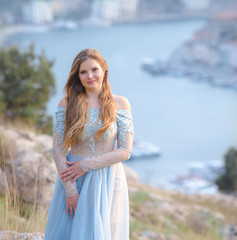 Fototapeta na wymiar A girl with long hair on the beach. She's wearing a beautiful blue dress . The bride of the sea. In the background view of the Bay, sea and city. Soft focus