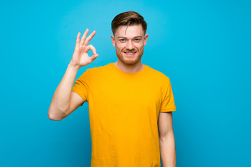 Redhead man over blue wall showing ok sign with fingers