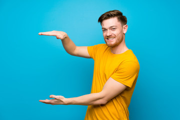 Redhead man over blue wall holding copyspace to insert an ad