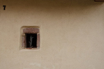 A small window with an old protective rod on a large wall of an old building. The cream-colored wall is covered with plaster. Background. Texture. Alsace. France. There is a number 7 on the wall.