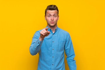 Blonde man over isolated yellow wall surprised and pointing front