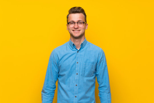 Blonde man over isolated yellow wall with glasses and happy