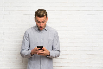 Blonde man over white brick wall surprised and sending a message