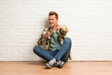 Fototapeta na wymiar Blonde man sitting on the floor pointing to the front and smiling