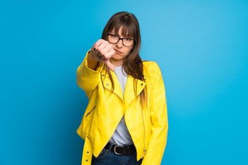 Young woman with yellow jacket on blue background showing thumb down sign with negative expression