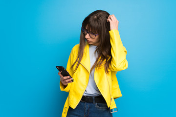 Young woman with yellow jacket on blue background with a mobile and thinking